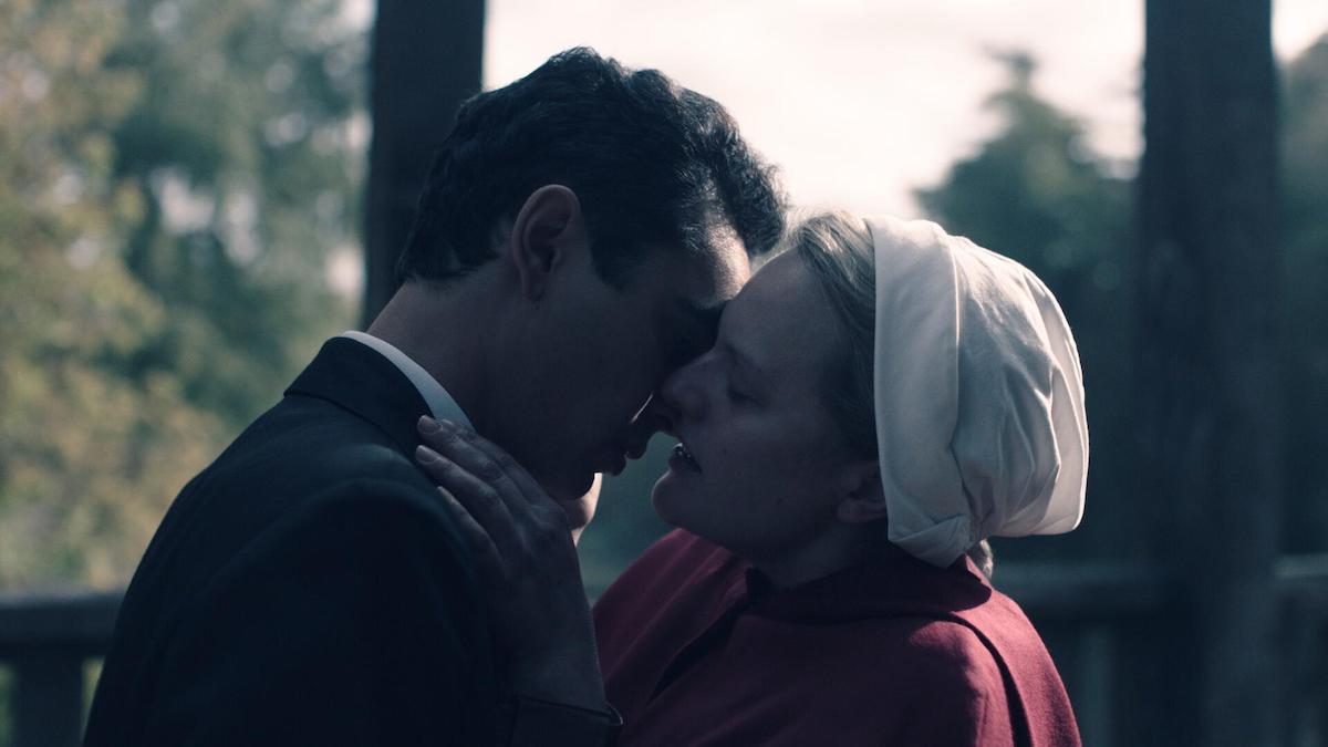 Elisabeth Moss and Max Minghella in 'The Handmaid's Tale' Season 4 Episode 2, 'Nightshade.' June stages an attack on a group of commanders in the episode but pays a price for it later. She's reunited with Nick in this photo. She's wearing her red Handmaid uniform and white bonnet as she leans in to kiss Nick, who's wearing a black suit. They're on a wooden bridge in daytime with trees surrounding.