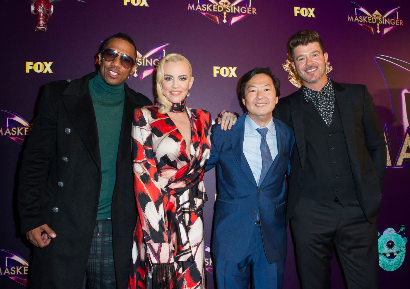 The host and judges panel of 'The Masked Singer' dressed professionally in front of a purple background with the logo