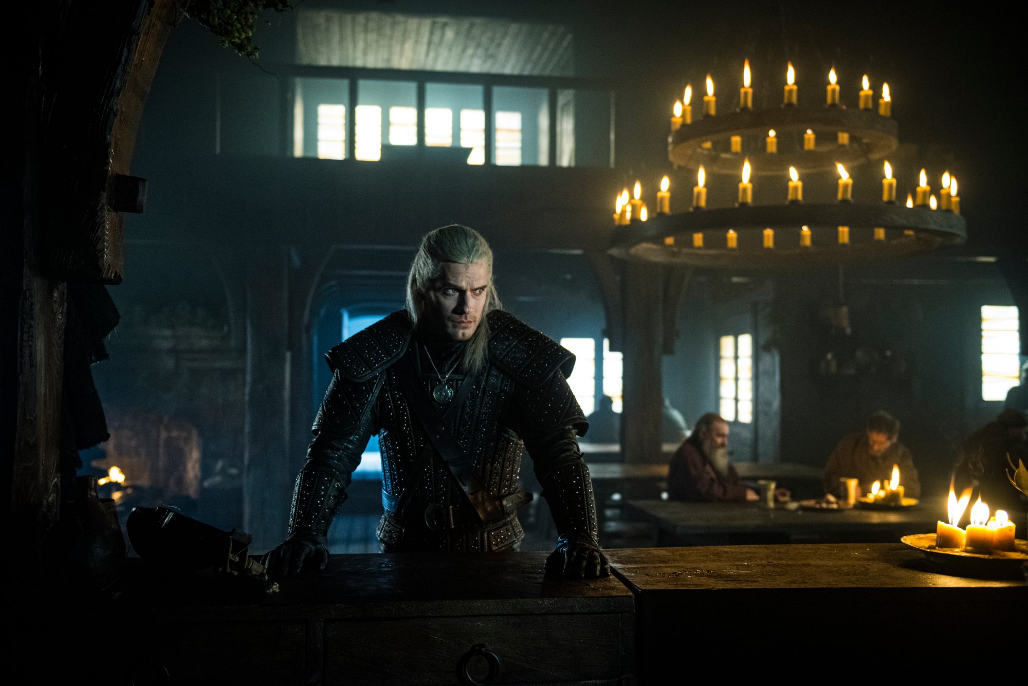 The Witcher star Henry Cavill as Geralt of Rivia. He stands wearing black leather and leaning on a table. Behind him are men eating and a chandelier. This takes place after his bathtub scene in season 1.