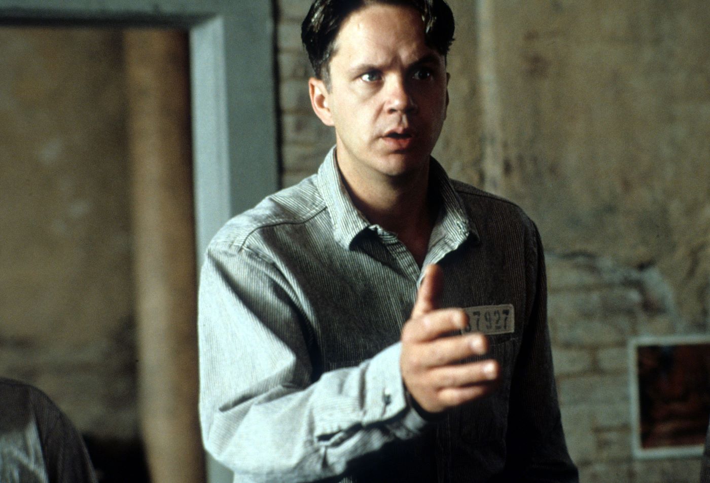 Tim Robbins in 'The Shawshank Redemption' dressed in his prison uniform in a stone room with his hand offered for a handshake.