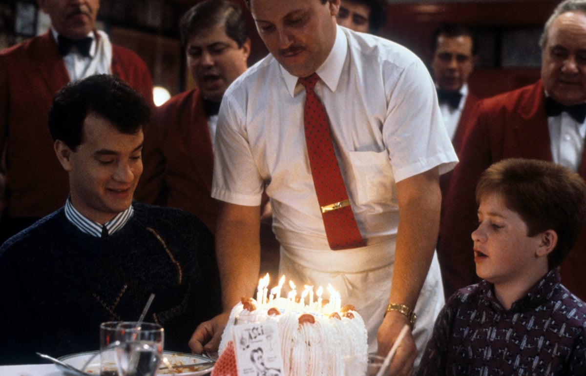 Tom Hanks sitting at a dining table as a birthday cake is being delivered in 'Big'