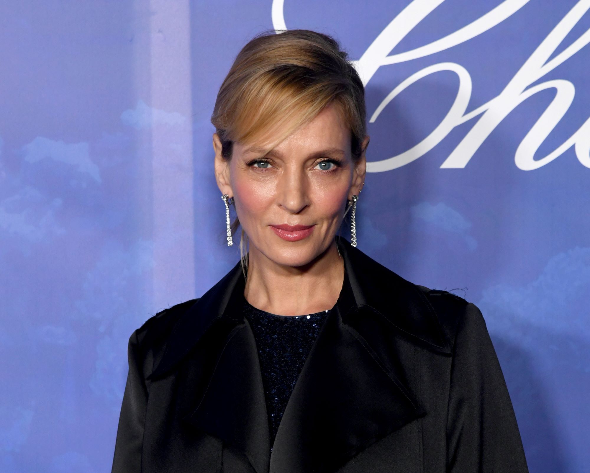 Uma Thurman wearing a black shirt with a black blazer in front of a blue background with cursive white writing.