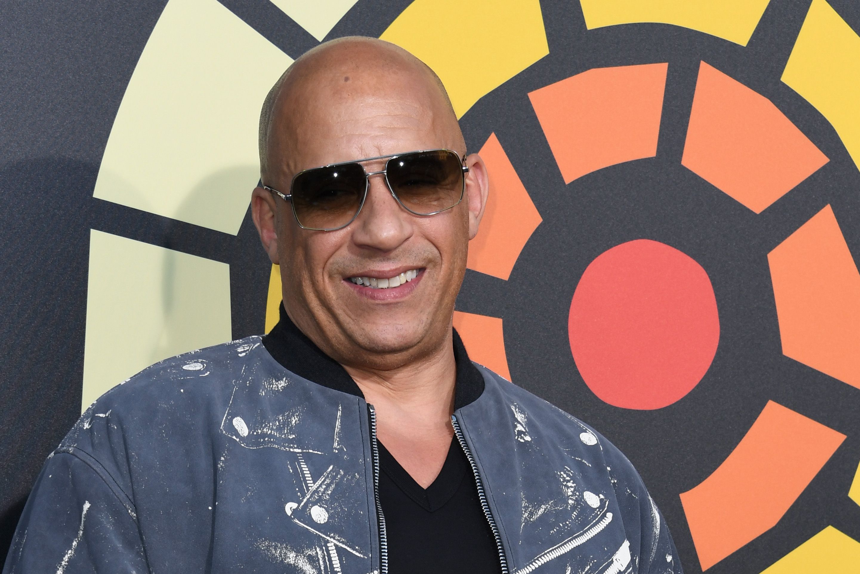 Vin Diesel smiling wearing sunglasses in front of an orange and yellow background.
