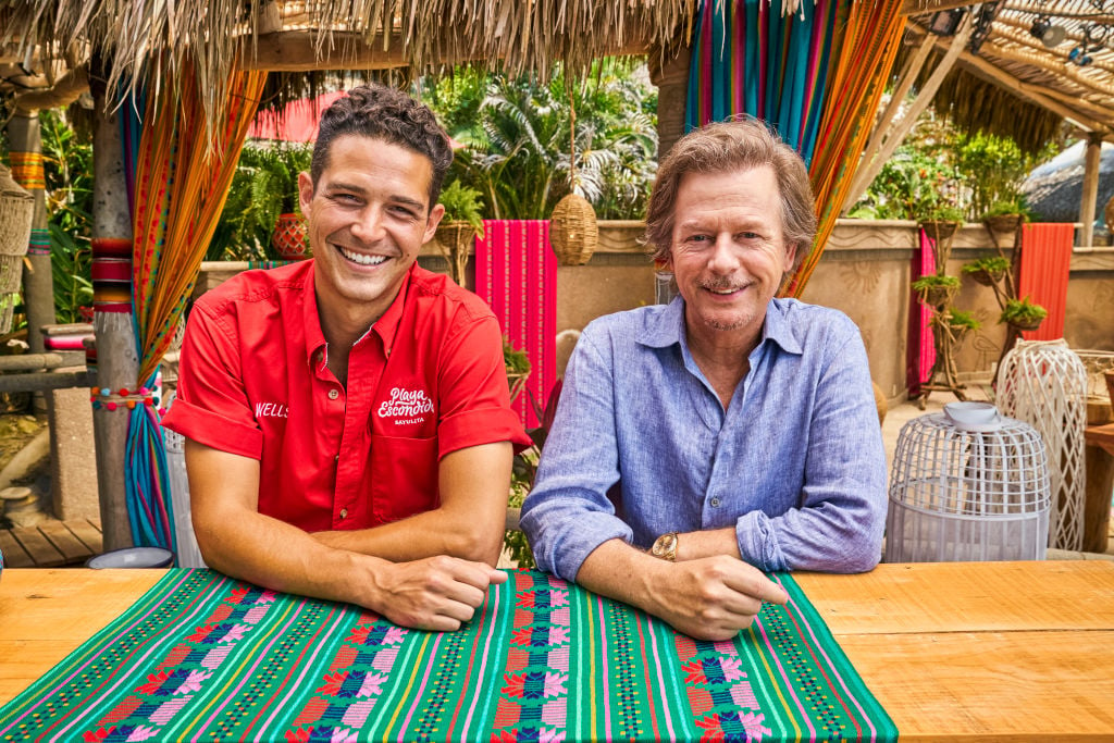 Wells Adams and David Spade pose for the camera, sitting side-by-side with smiles. Wells is dressed in a red shirt and David is wearing a light blue shirt.