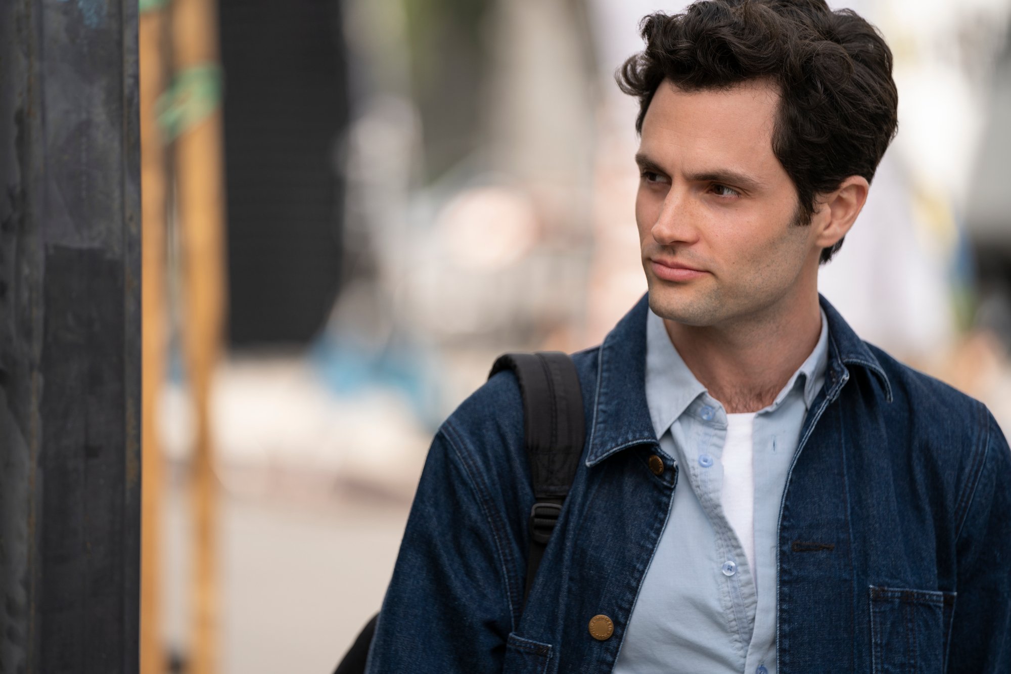 Penn Badgley as Joe in 'You' Season 2. He's wearing a blue denim jacket and blue button-up shirt and looking at something off-screen. The season 3 teaser trailer sees him becoming a dad.