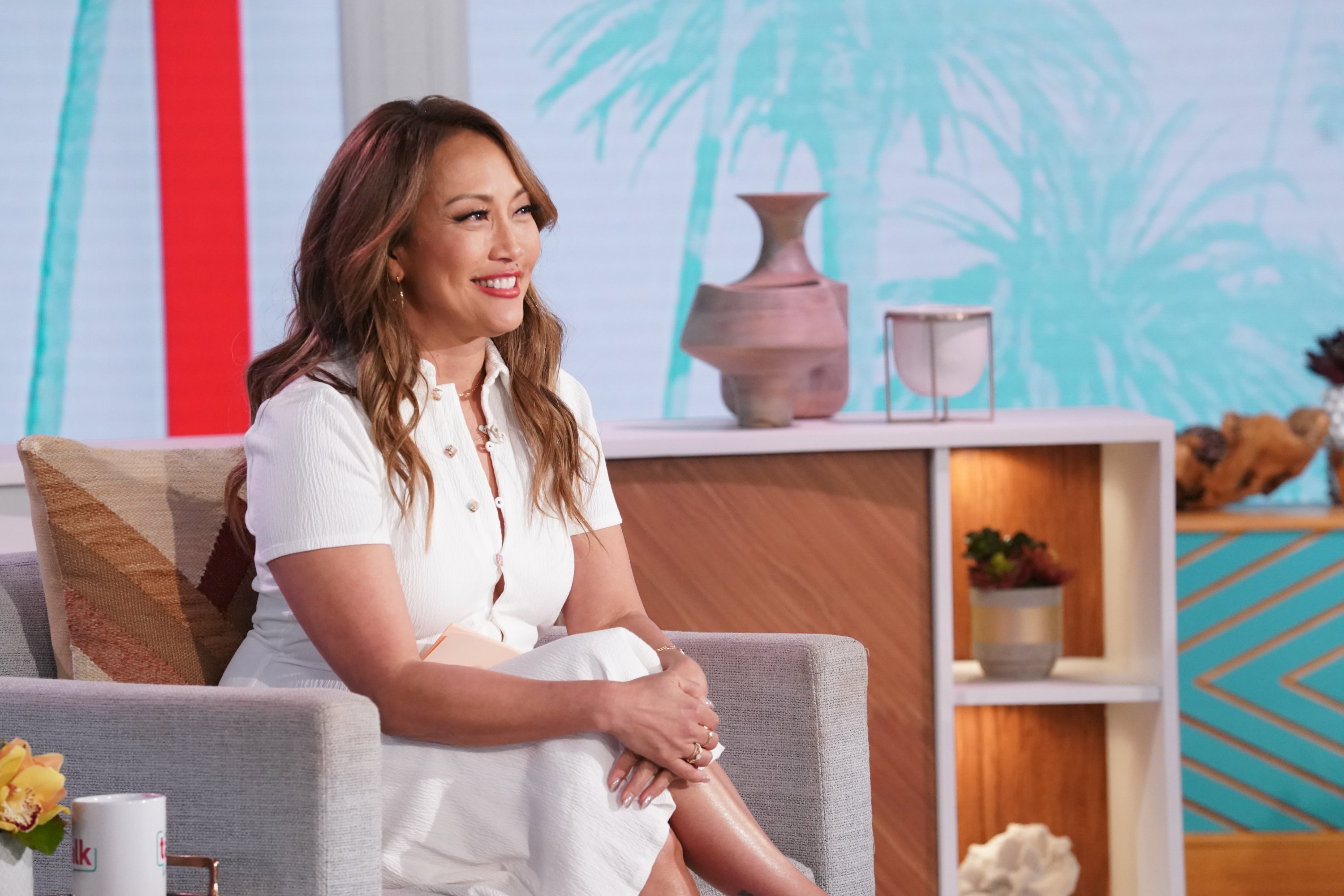 Carrie Ann Inaba smiling and sitting in her co-host chair wearing a white dress on the set of 'The Talk'