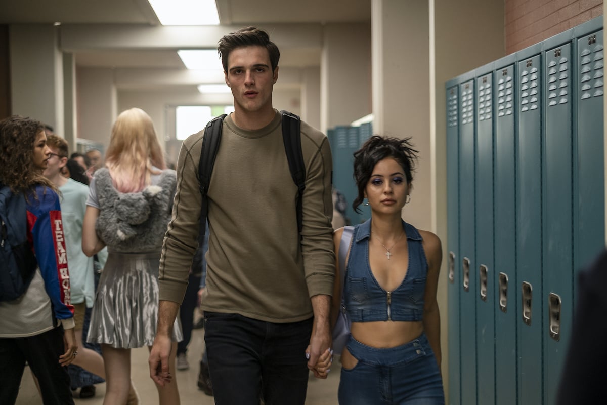 Jacob Elordi and Alexa Demie as Nate and Maddy in 'Euphoria' Season 1. The actors hold hands while walking down a high school hallway. Blue lockers line the halls. Elordi wears a tan long-sleeved shirt and black jeans and Demie wears a denim crop top with matching pants.