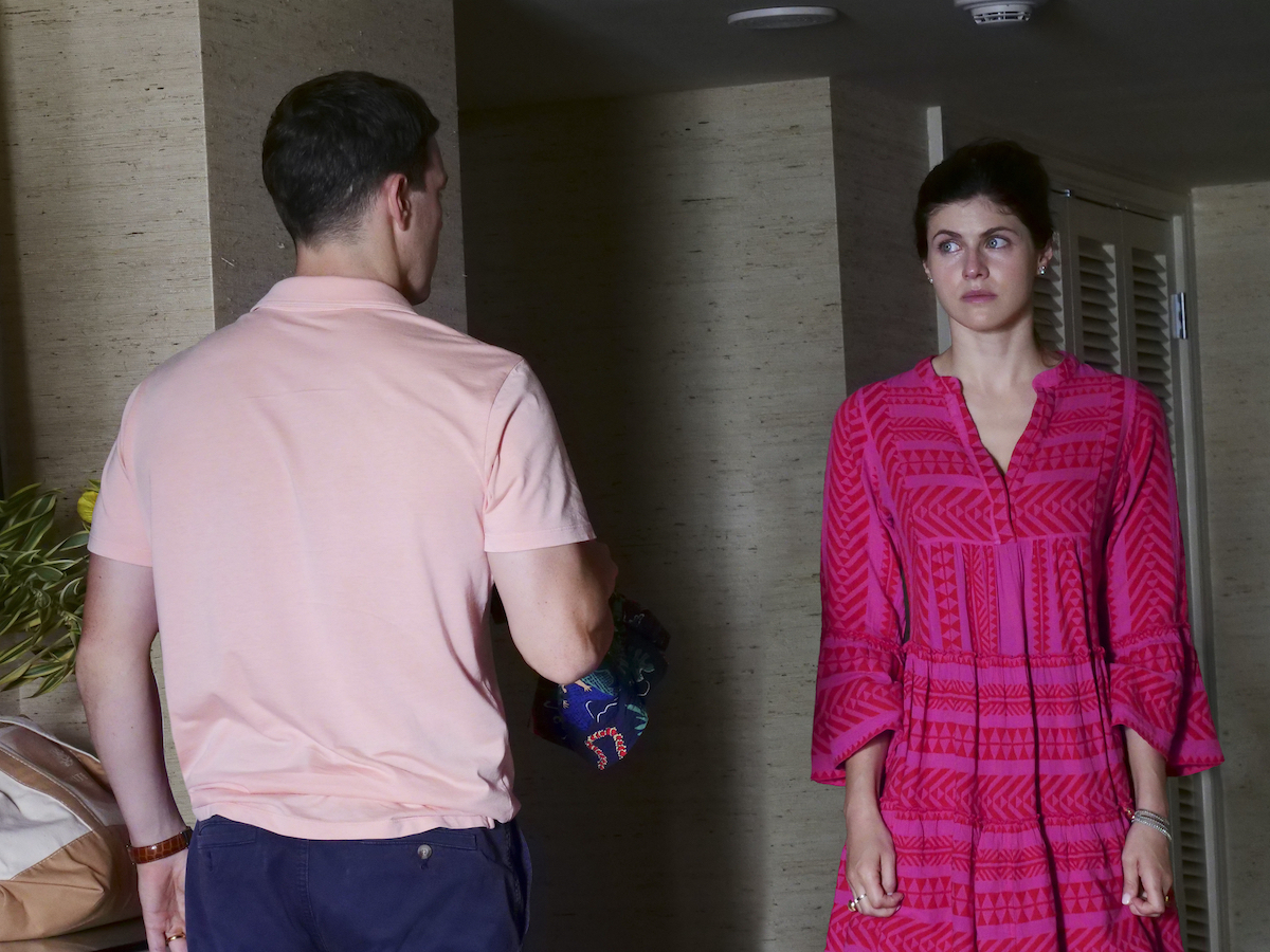 Jake Lacy and Alexandra Daddario in 'The White Lotus' Season 1 Episode 6. In 'The White Lotus' finale, Rachel Patton talks about making a 'faustian bargain' with her marriage to Shane. In this photo, Daddario wears a hot pink, patterned, long-sleeved dress with her hair up in a bun. She gives Lacy a tense look in a hotel room. Lacy wears navy blue shorts and a light pink shirt and his back is facing the camera. Sunlight's coming in from an off-camera window. The walls are beige and slightly striped.