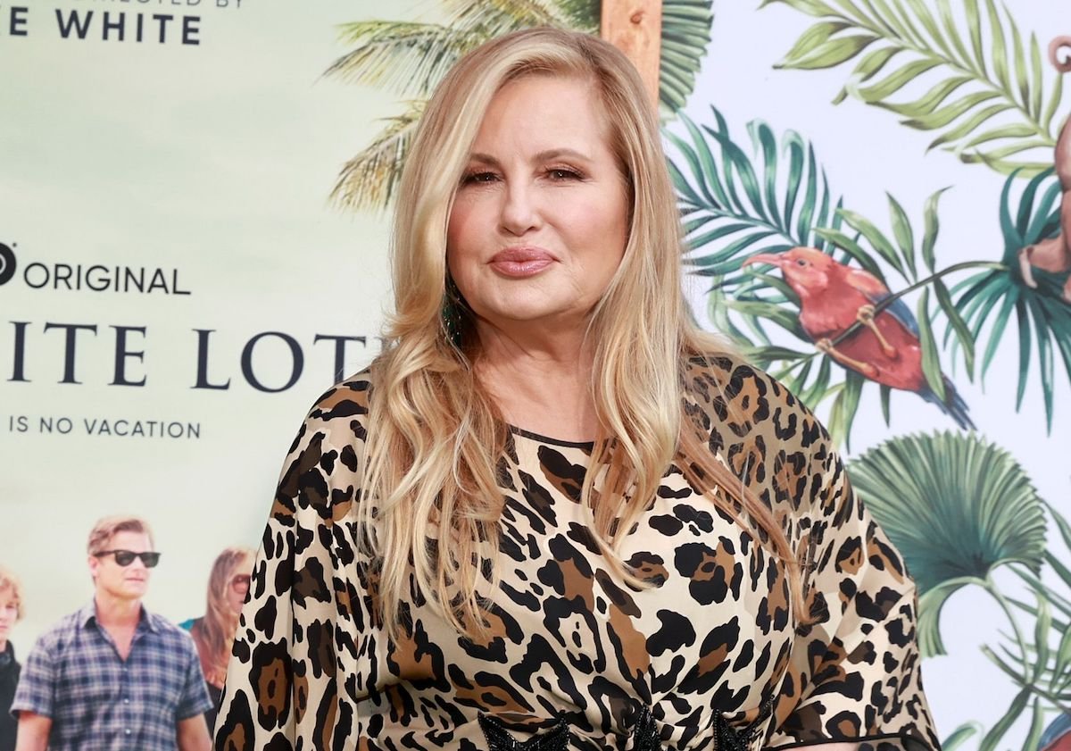 Jennifer Coolidge attends the Los Angeles premiere of HBO's 'The White Lotus.' She wears a long-sleeved leopard print dress, her long blonde hair is styled down, and she stands in front of a tropical backdrop that says 'The White Lotus' and shows a photo of the cast.