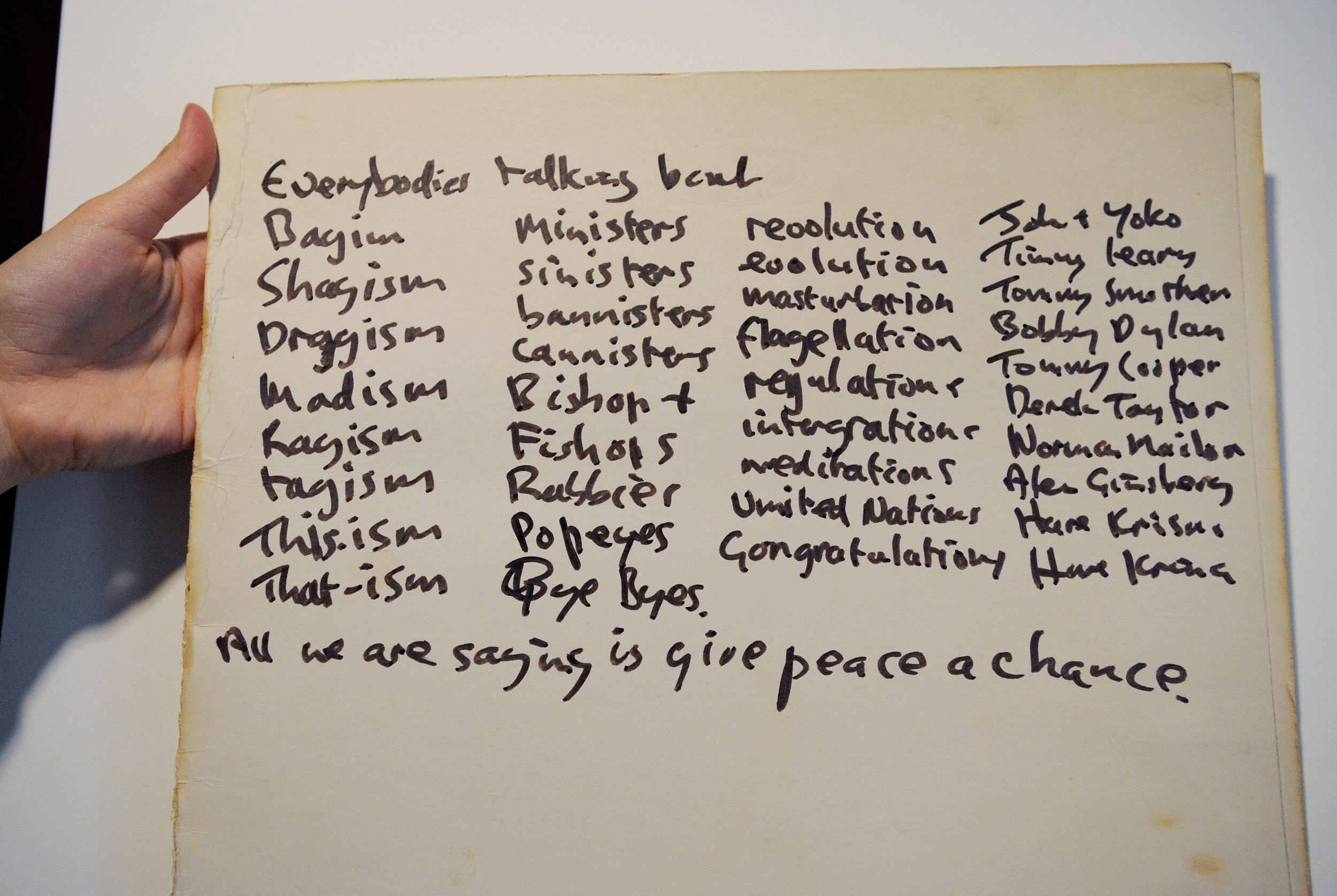 A hand holding John Lennon's handwritten lyrics for "Give Peace a Chance"