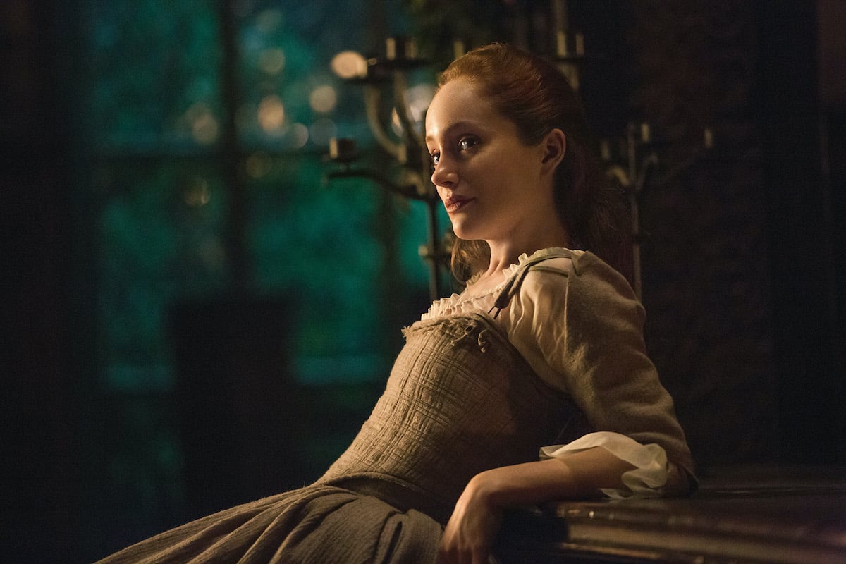 Lotte Verbeek as Geillis Duncan in 'Outlander' Season 1. Verbeek leans her arms and back against a wooden counter at nighttime. She wears a beige dress fitting the 1700s Scotland time period. There are large brass candlesticks behind her and large windows showing trees outside. 'Outlander's Geillis Duncan was featured in season 1, season 2, and season 3, and Verbeek says she could come back to the 'Outlander' cast in the future. Fans are hoping she will be part of the 'Outlander' Season 6 cast.