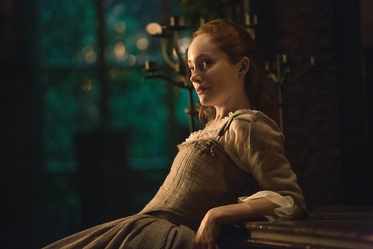 Lotte Verbeek as Geillis Duncan in 'Outlander' Season 1. Verbeek leans her arms and back against a wooden counter at nighttime. She wears a beige dress fitting the 1700s Scotland time period. There are large brass candlesticks behind her and large windows showing trees outside. Given Geillis' time traveling abilities, 'Outlander' fans wondered if Claire and Geillis are related. As it turns out, they are. And they're connected by another time traveler.