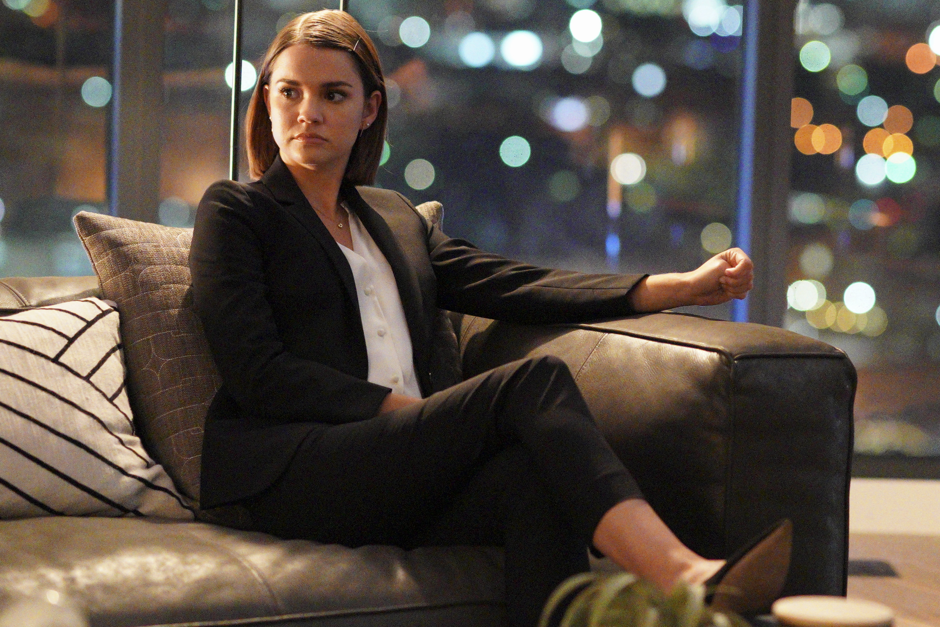 Maia Mitchell sits on a couch while wearing her lawyer suit on the set of 'Good Trouble.'