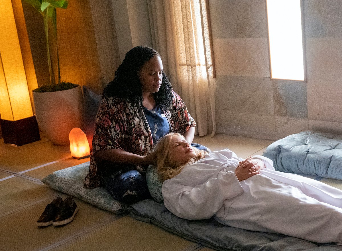 Natasha Rothwell and Jennifer Coolidge in HBO's 'The White Lotus.' Coolidge lays on her back on the floor while wearing a white robe and closing her eyes and holding her hands on her stomach as Rothwell massages her head. A Himalayan salt lamp is on the floor behind them. Rothwell sits on a blue, coral patterned pillow and Coolidge lays on a long, blue cushion.