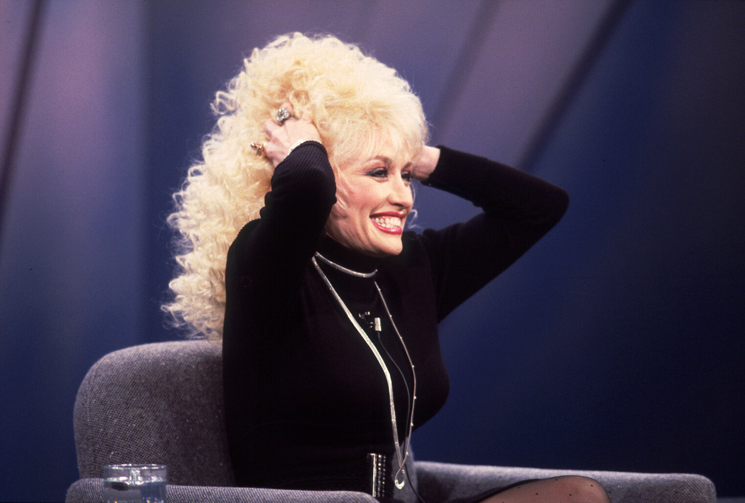 Dolly Parton sitting on the set of the Oprah Winfrey Show. Her hands are in her hair.