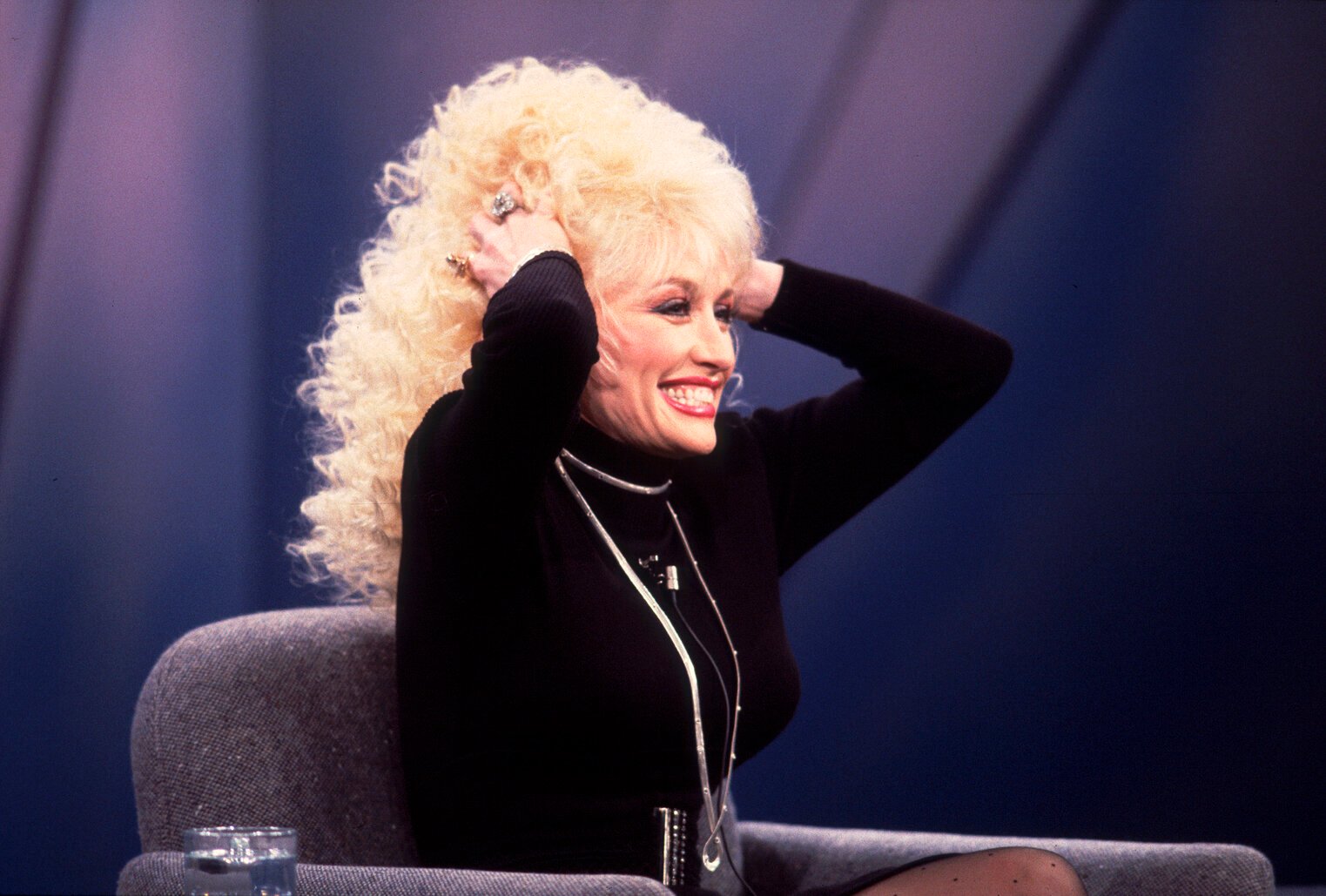 Dolly Parton sitting on the set of the Oprah Winfrey Show. Her hands are in her hair.