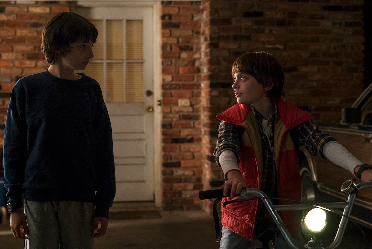 'Stranger Things' characters Mike Wheeler (Finn Wolfhard), in a blue sweatshirt and gray sweatpants and talks to Will Byers (Noah Schnapp) in a red puffy vest sitting on a bikee.