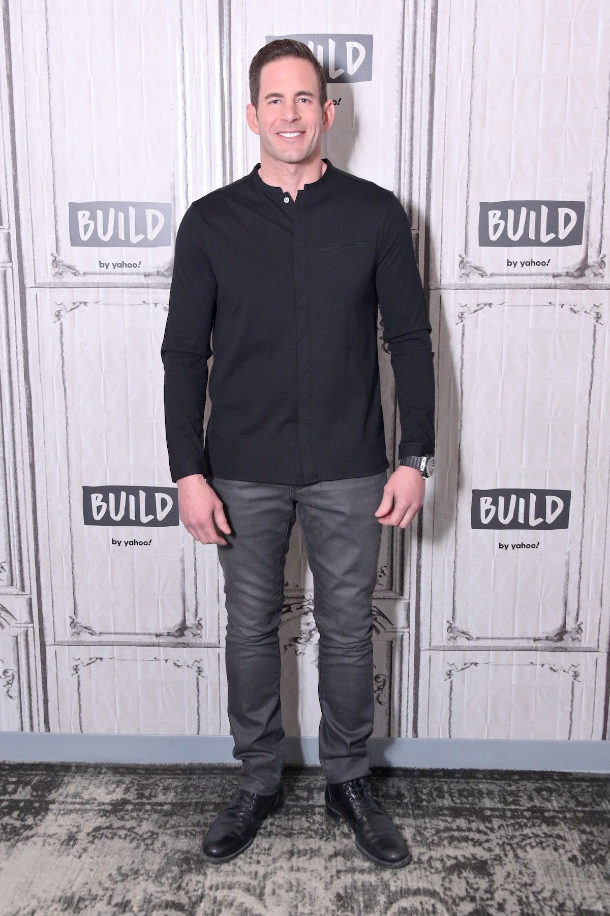 Tarek El Moussa posing in front of a step and repeat in a black shirt with the top button unbuttoned and gray slacks