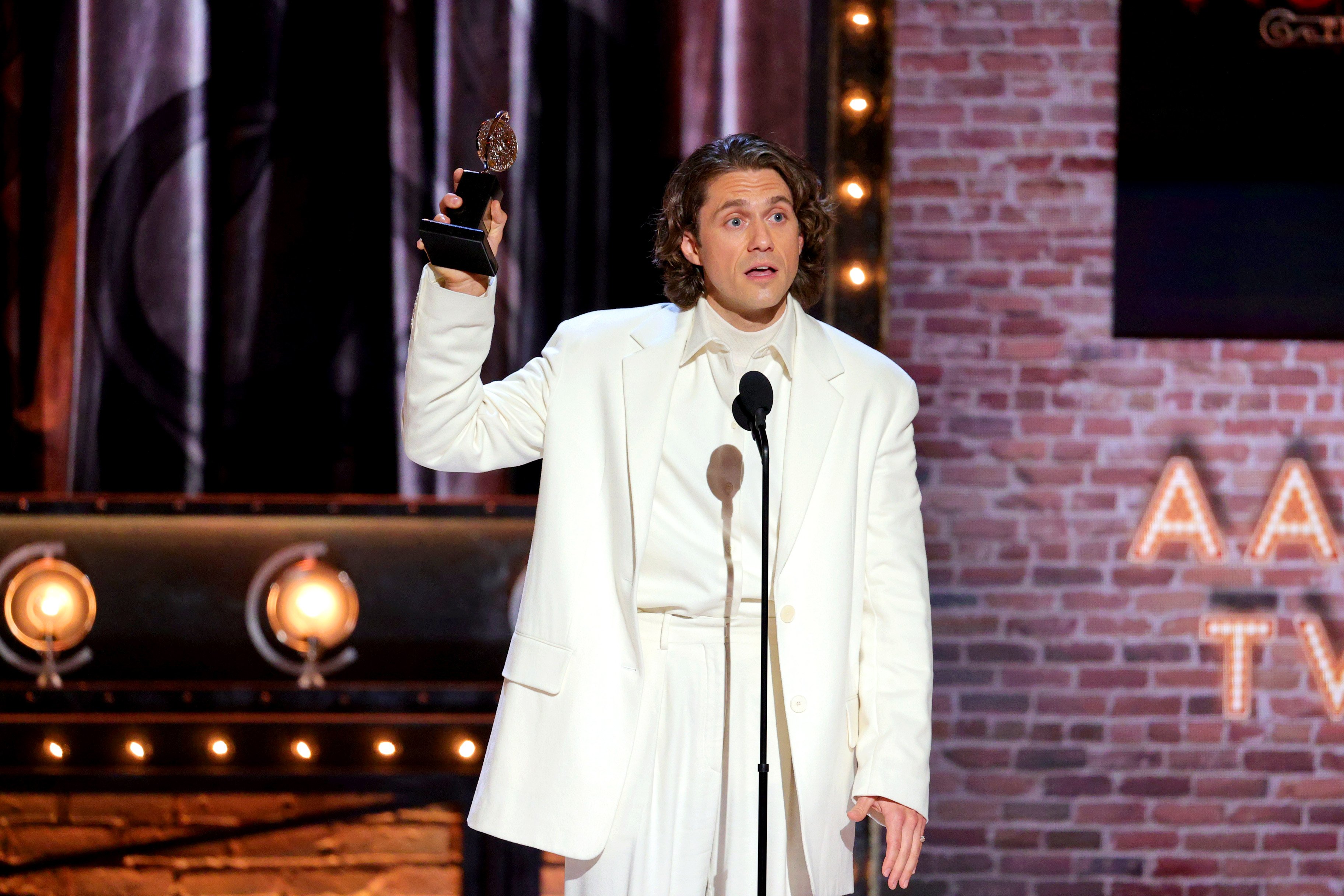Aaron Tveit holds up a Tony Award while wearing a white suit