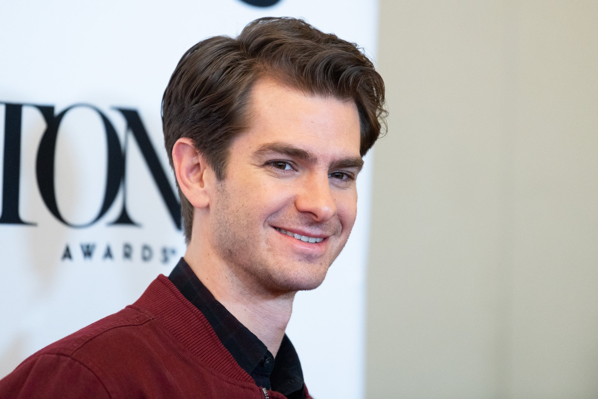 'The Amazing Spider-Man' actor Andrew Garfield. He's wearing a red jacket and black shirt, and he's smiling at the camera. His brown hair is slicked to the side.
