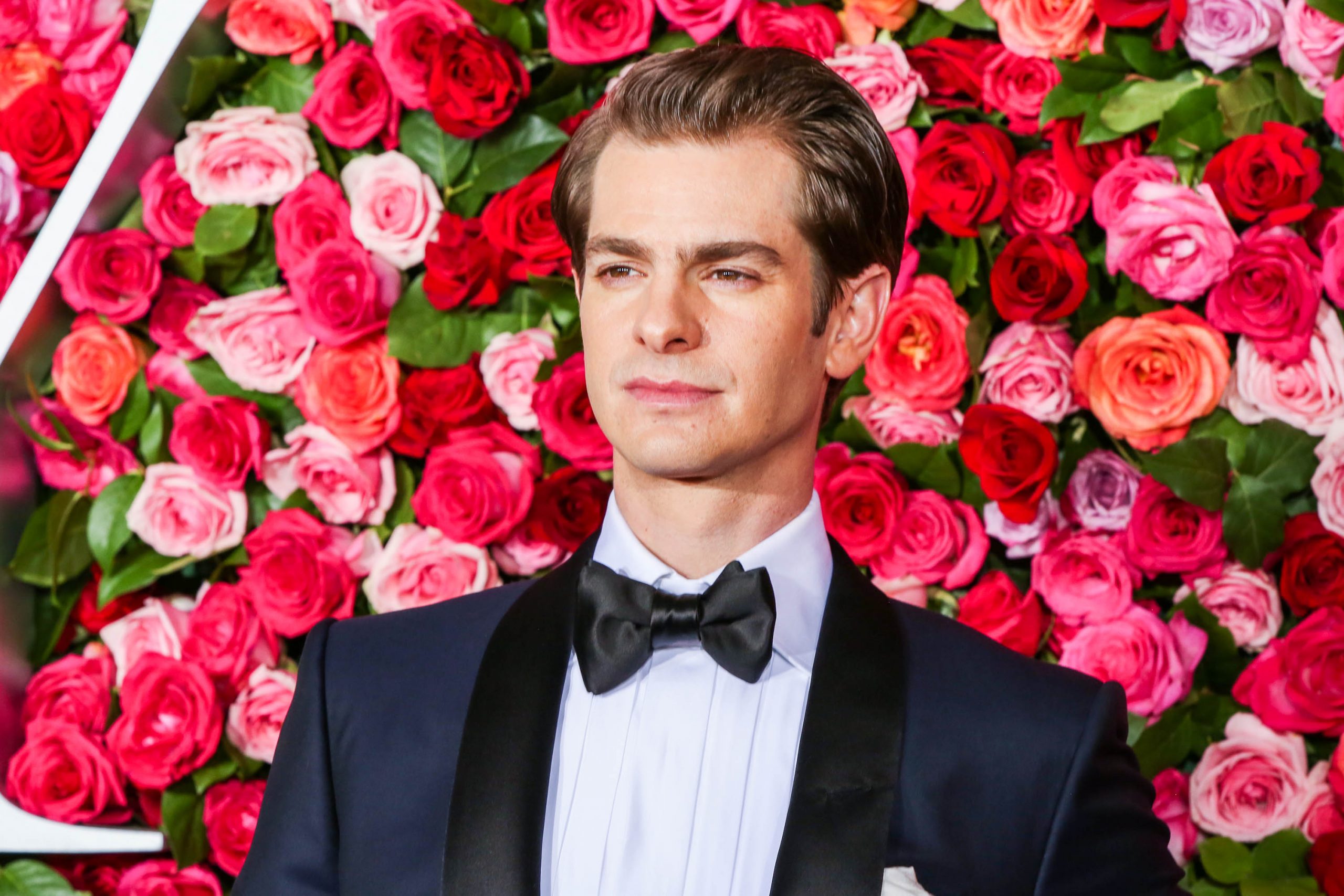 Andrew Garfield posting in a white shirt and blue tuxedo with a bow-tie. He is standing in front of a rose background at the 72nd Annual Tony Awards in 2018.