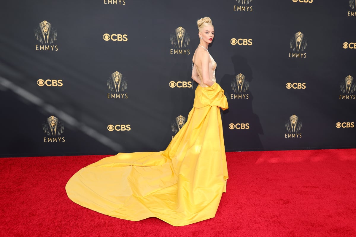 Anya Taylor-Joy wears a form-fitting, backless gown with a long yellow cape on the red carpet. Her hair is in an intricate up-do. 