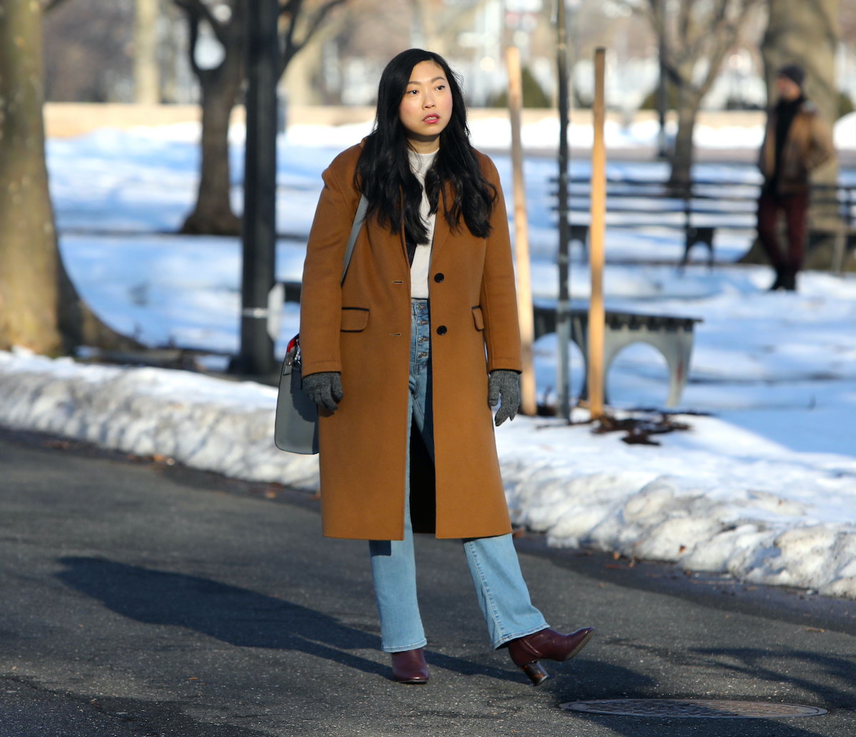 Awkwafina wears a brown coat and jeans as she looks on while filming a scene from 'Awkwafina Is Nora From Queens' in 2021