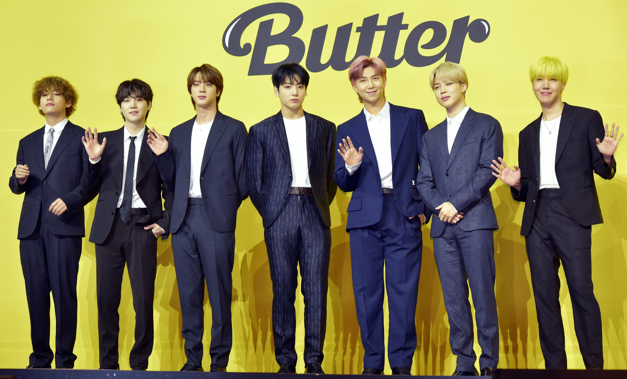 V, Suga, Jin, Jungkook, RM, Jimin, and J-Hope stand in front of a yellow backdrop with the word 'Butter'