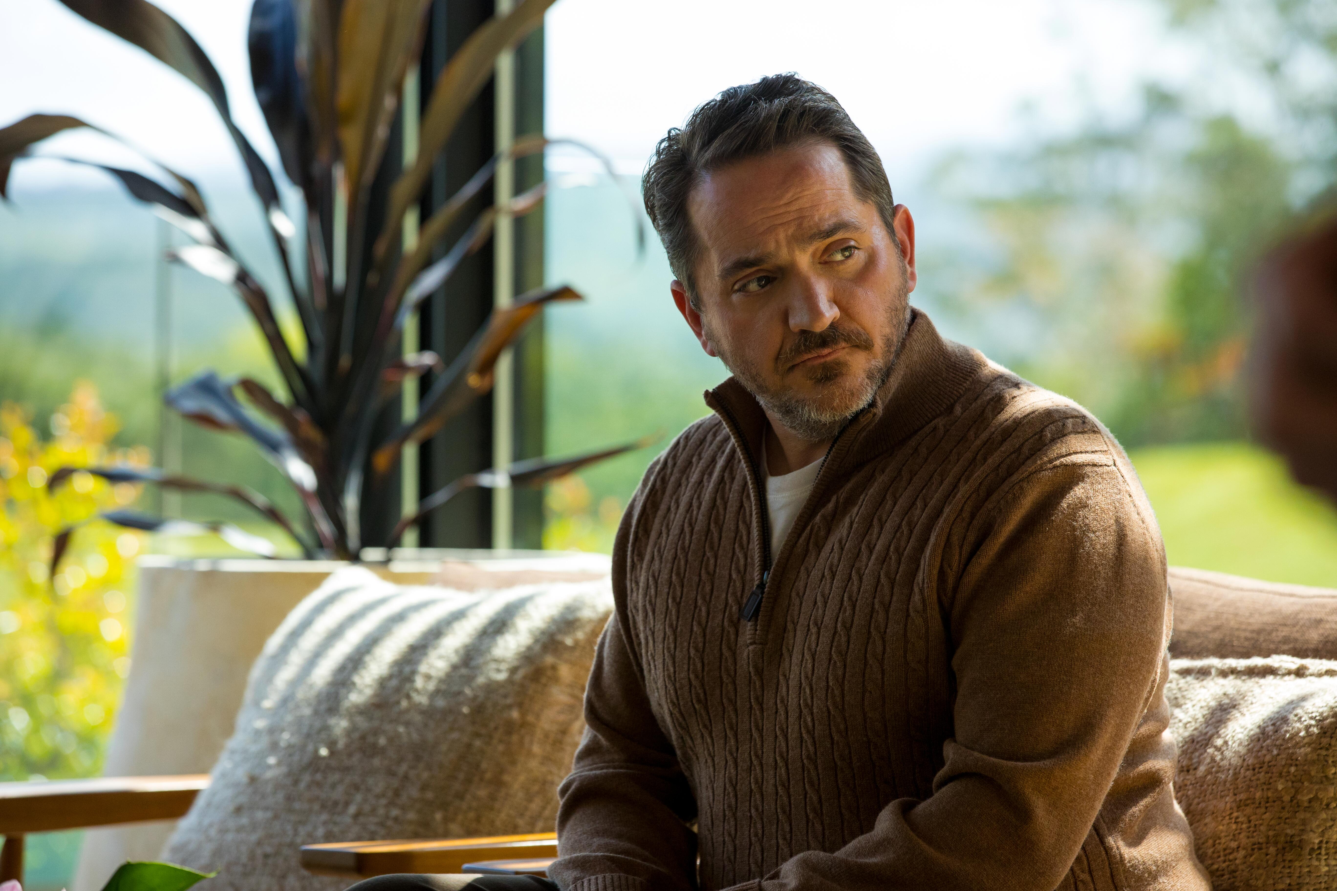 Ben Falcone as Paul Drabble sits on the couch at Tranquillum House.