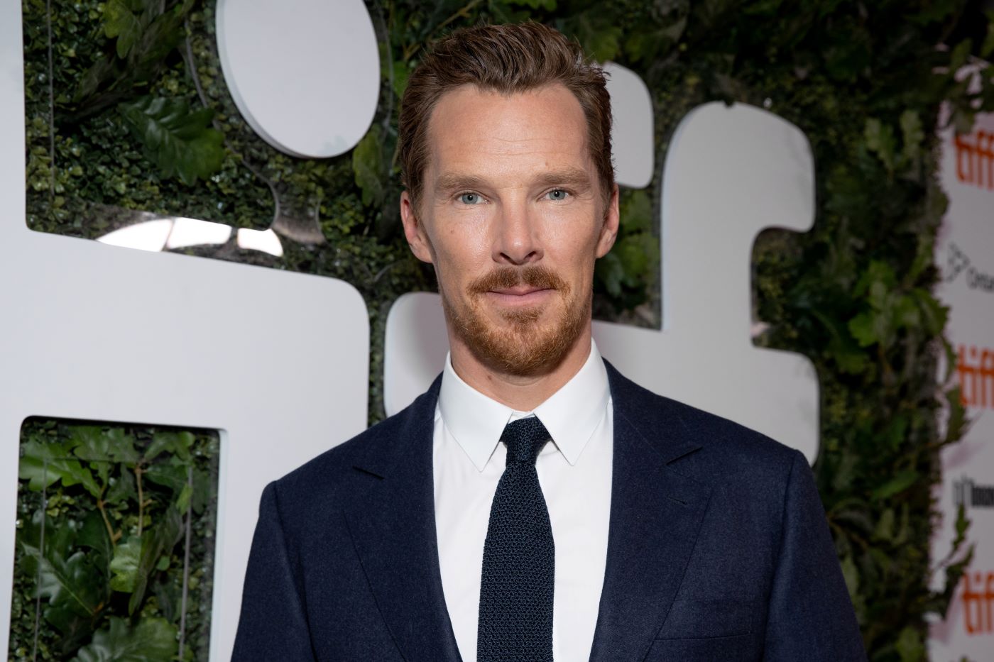 Benedict Cumberbatch is wearing a black suit standing in front of a background with green leaves and white writing.