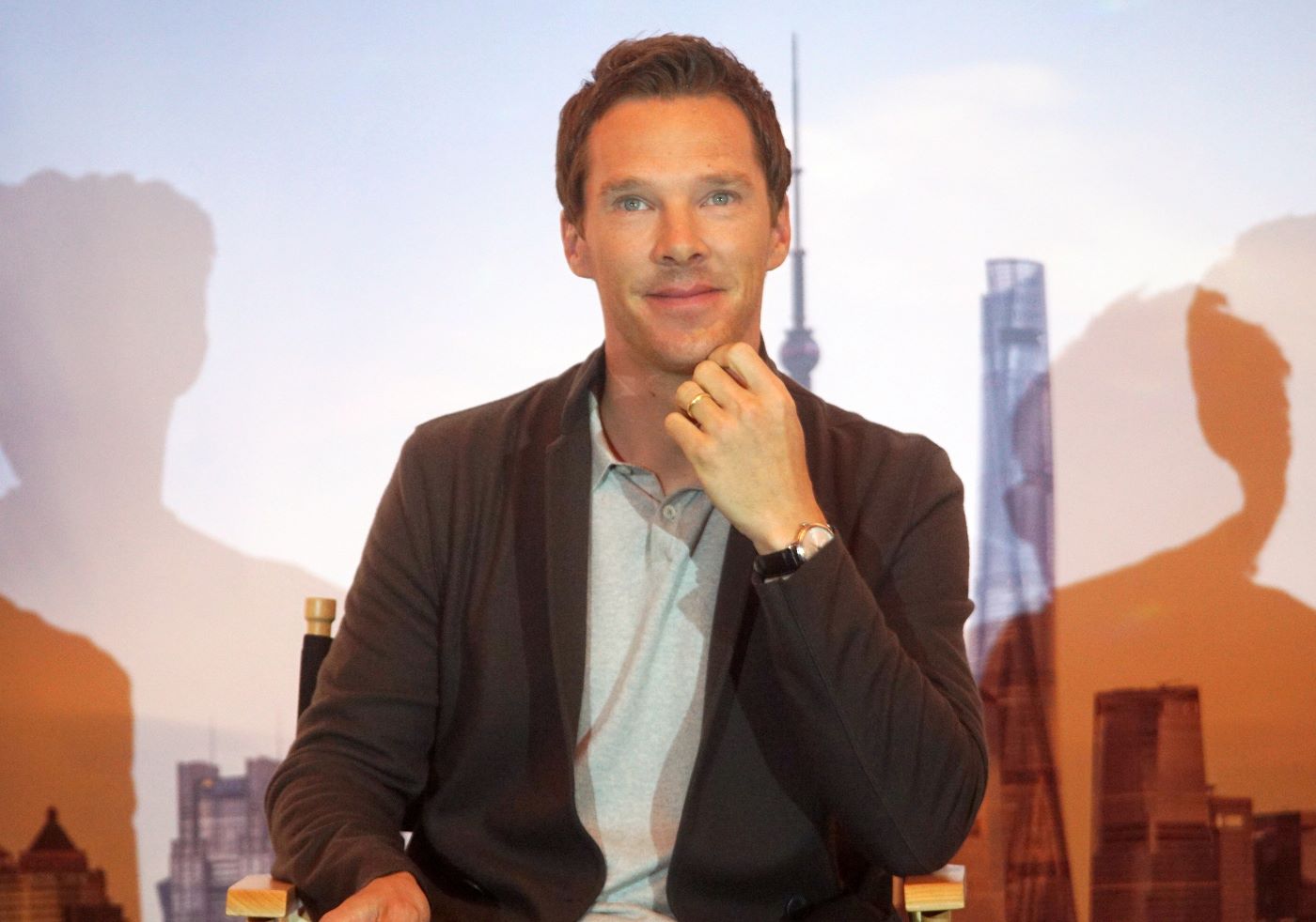 Benedict Cumberbatch from 'Dr. Strange' and 'Spider-Man: No Way Home' sitting in front of a picture of a city skyline wearing a brown cardigan and a gray polo.