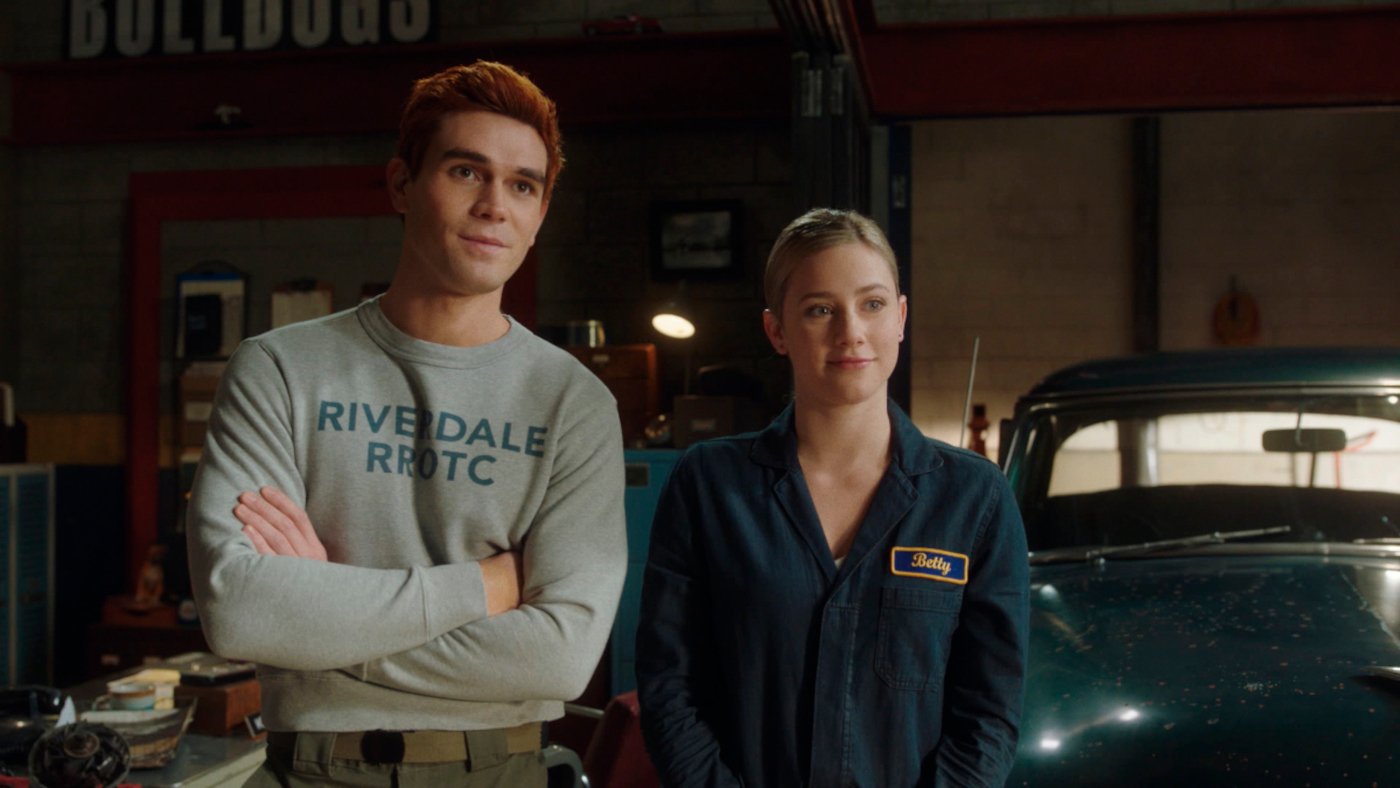 KJ Apa and Lili Reinhart as Archie and Betty in 'Riverdale' Season 5. They're standing next to one another and smiling. His arms are crossed.