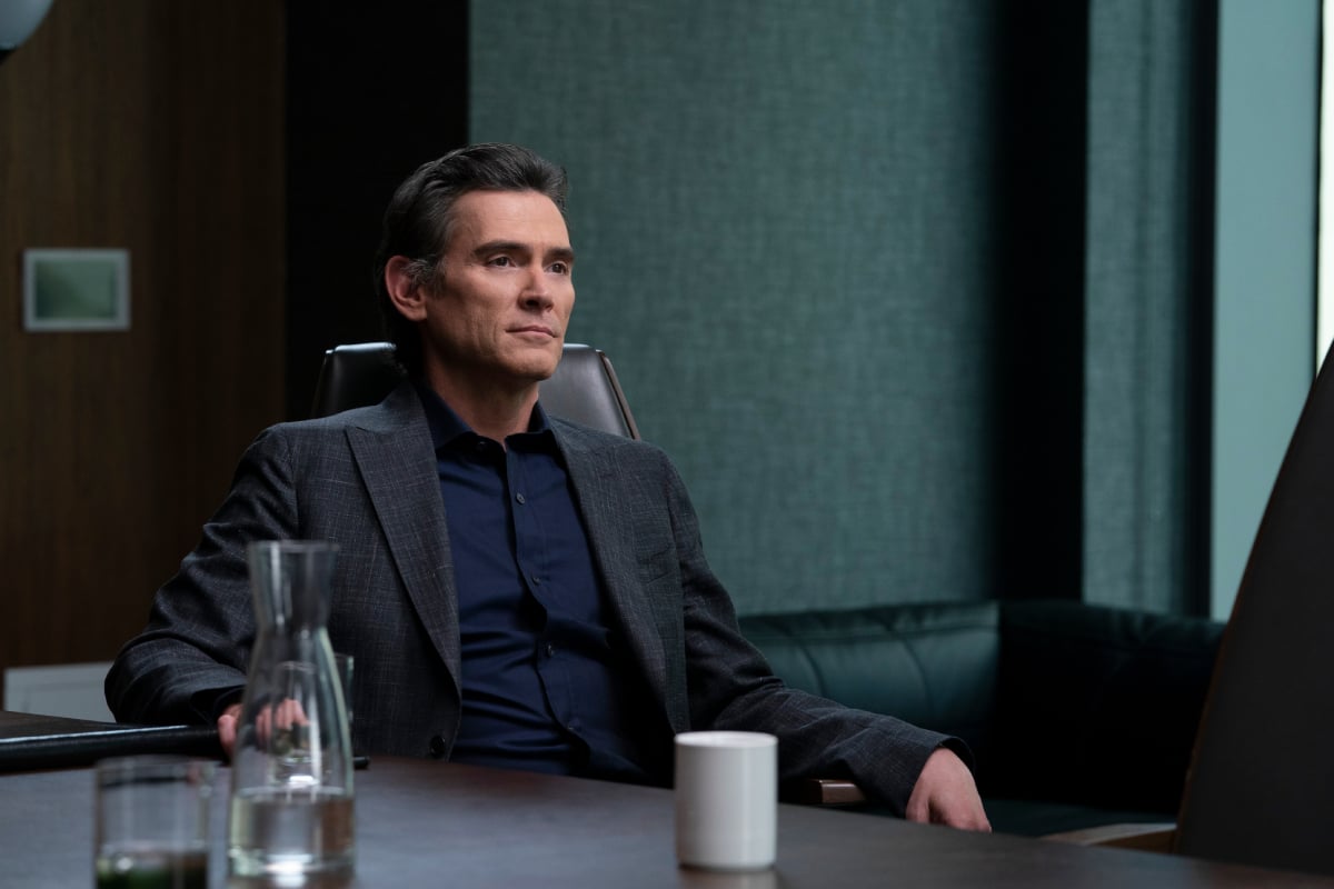 Billy Crudup sits in a chair with a neutral expression in 'The Morning Show.' He is wearing a dark blue button-up and a suit jacket.