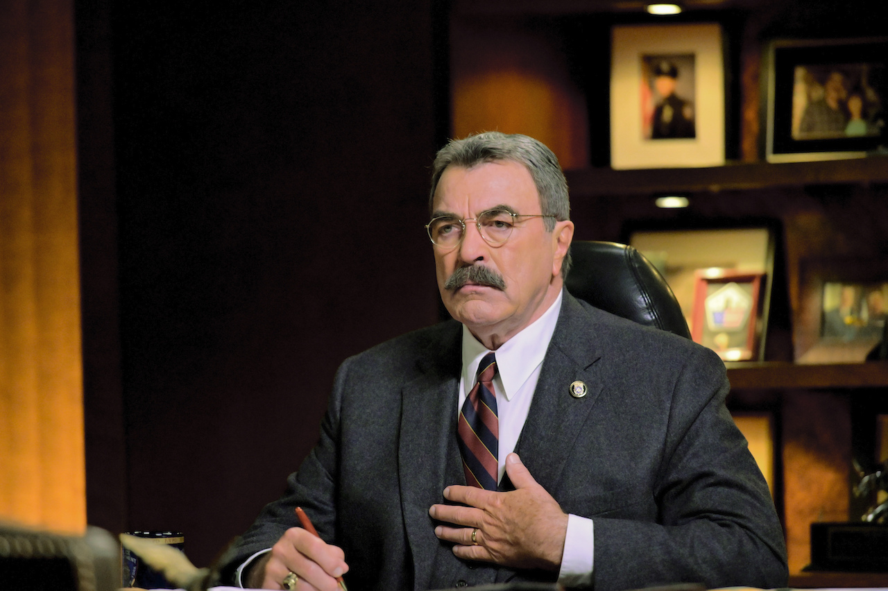 Tom Selleck as Frank Reagan sits at his desk wearing a suit on 'Blue Bloods'