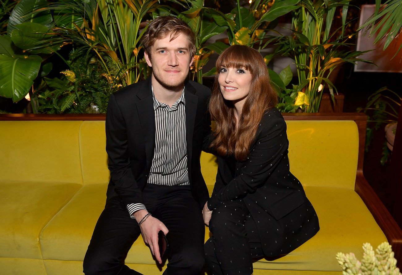 Bo Burnham and Lorene Scafaria wear black outfits and sit on a yellow couch.