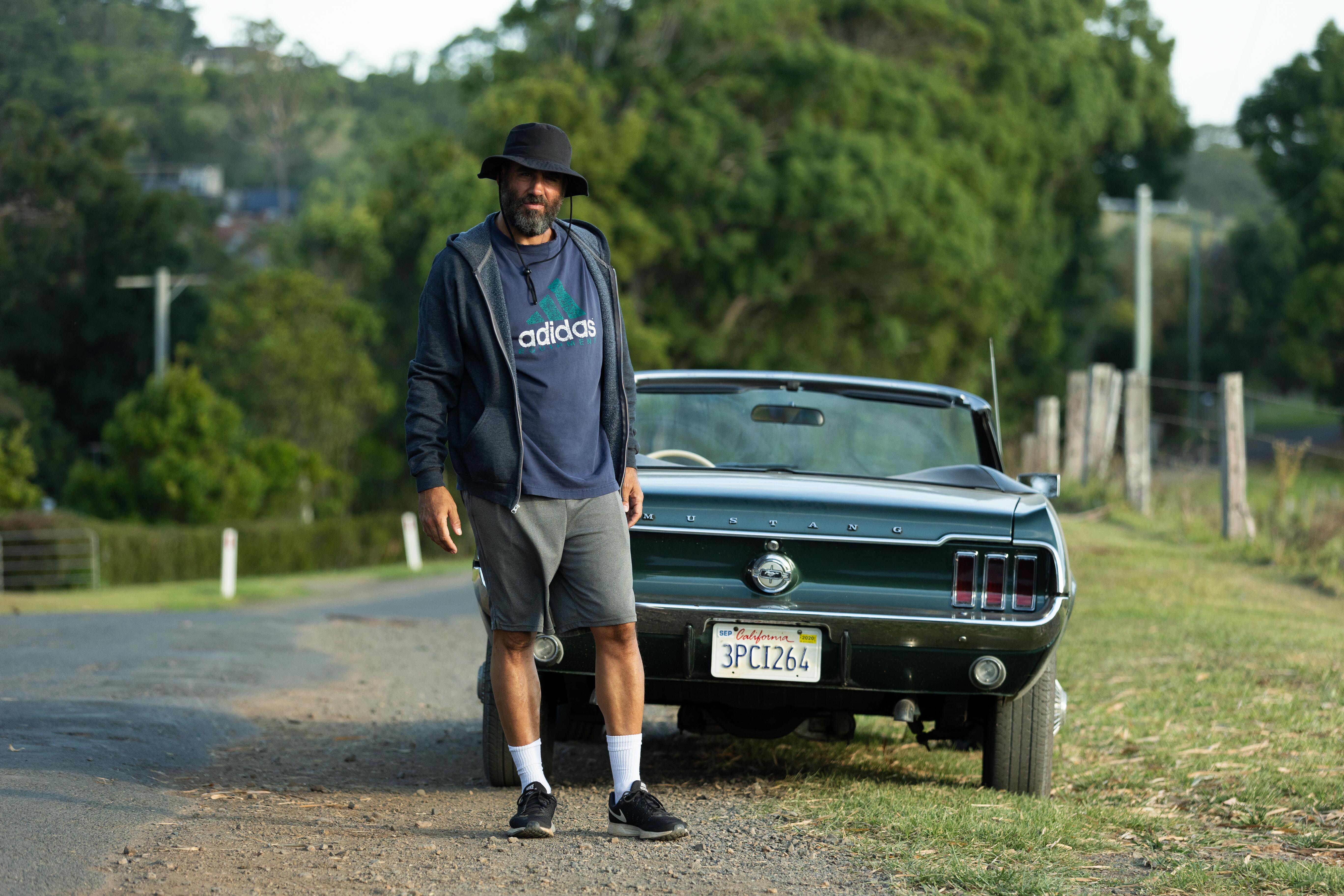 Tony Hogburn stands outside his car wearing an Adidas t-shirt and wide-brimmed hat. 