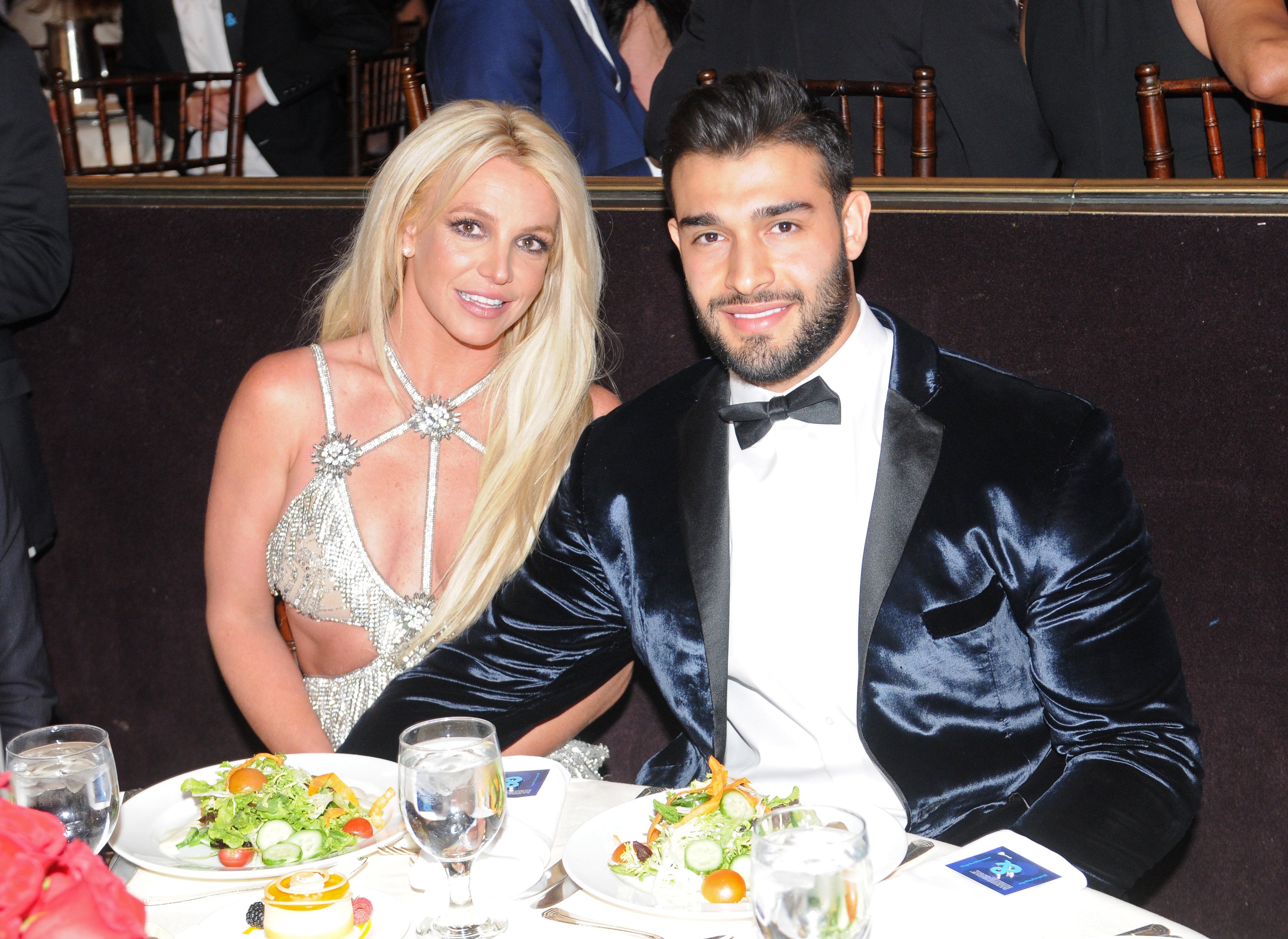 Britney Spears and Sam Asghari smiling and holding hands while sitting down.