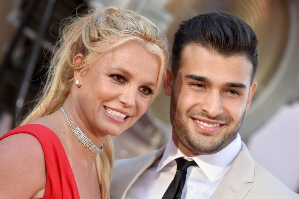 Britney Spears and Sam Asghari walk the red carpet. Spears is in a red dress while Asghari wears a tan suit.