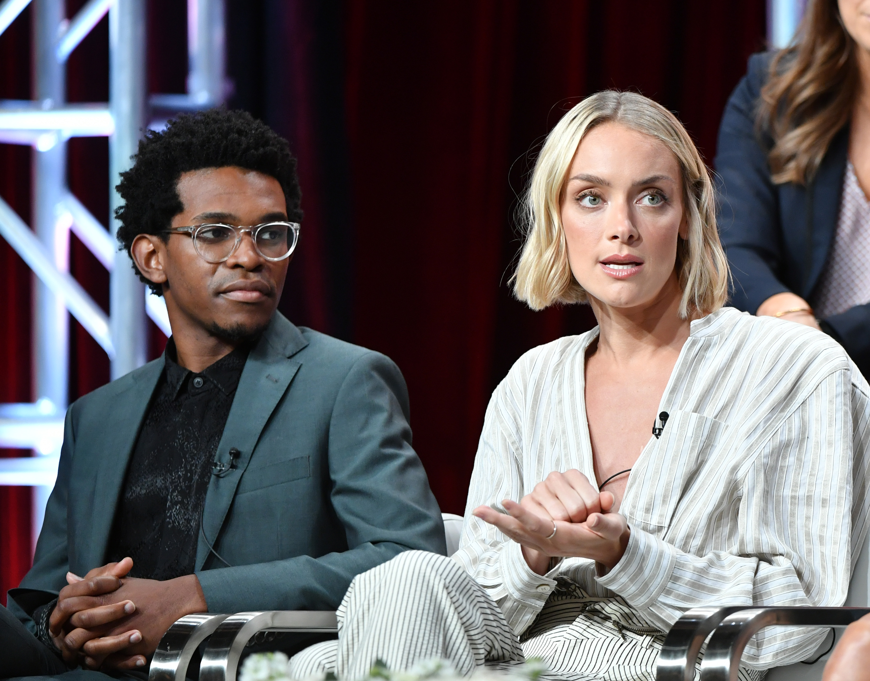 'Batwoman' stars Camrus Johnson, dressed in a gray suit, and Rachel Skarsten, dressed in a white striped shirt, speak onstage.