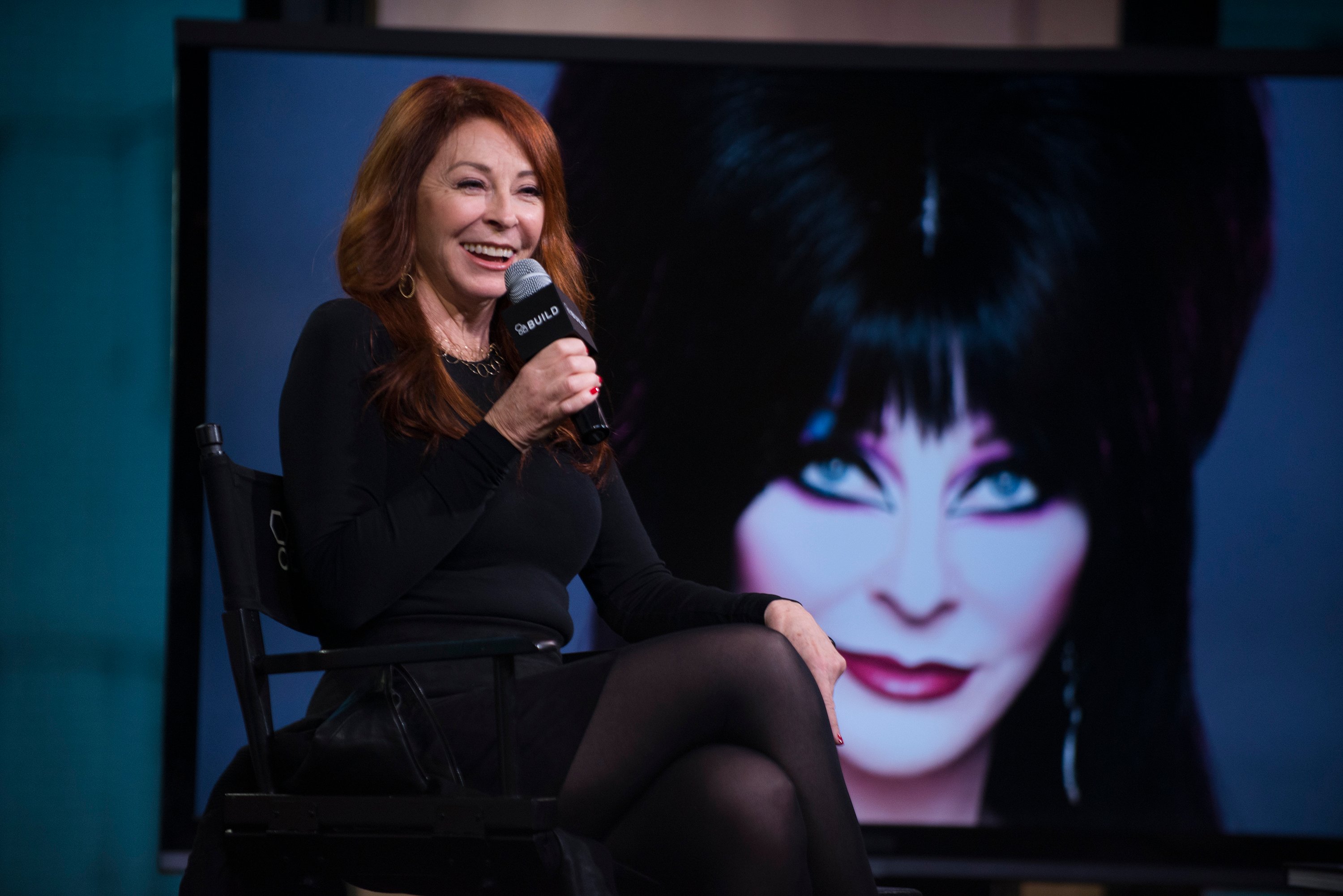 Cassandra Peterson, in a black dress, sitting in front of her Elvira, Mistress of the Dark poster, at the AOL Build Series in 2016.
