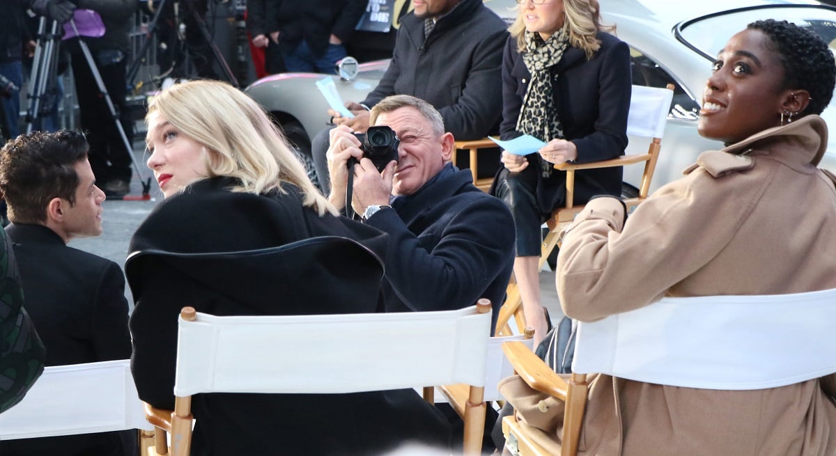 Lea Seydoux on the left and Lashana Lynch on the right both sitting on chairs looking up at something that Daniel Craig is taking a picture of. 