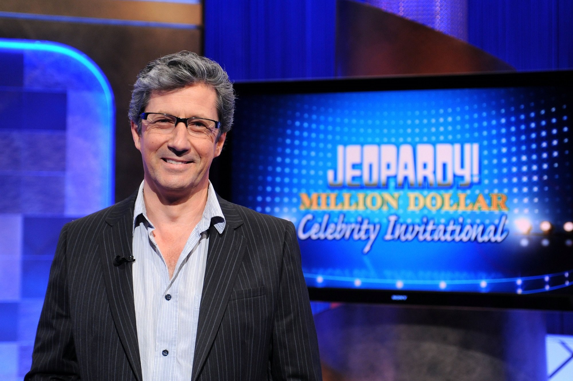 Charles Shaughnessy of Days of Our Lives and General Hospitalin a blue tailored suit with the Jeopardy! logo behind him