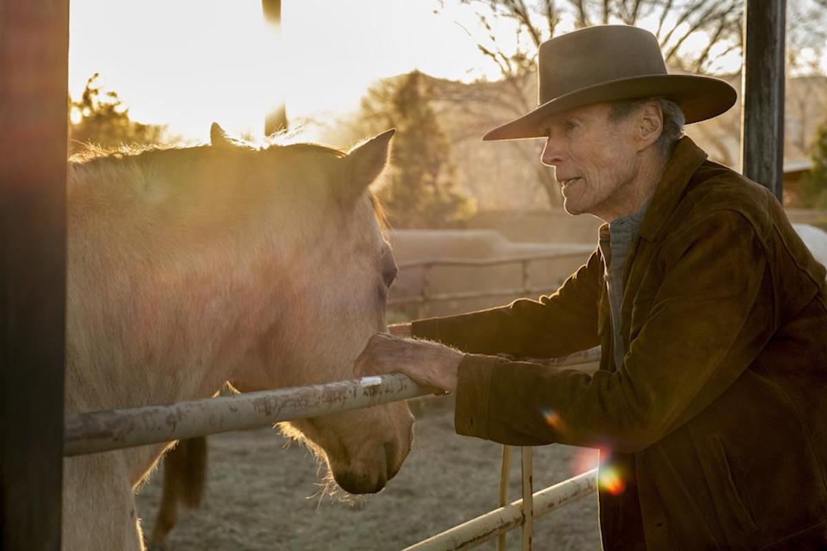 Clint Eastwood on the set of 'Cry Macho.' He wears a cowboy hat and brown suede jacket while leaning against a fence. A blonde-haired horse is on the other side of the fence and the sun is shining bright behind them. 'Cry Macho,' out Sept. 17, marks Eastwood's first western since 1992's 'Unforgiven,' and it required him to ride a horse for the first time since filming 'Unforgiven' three decades ago.