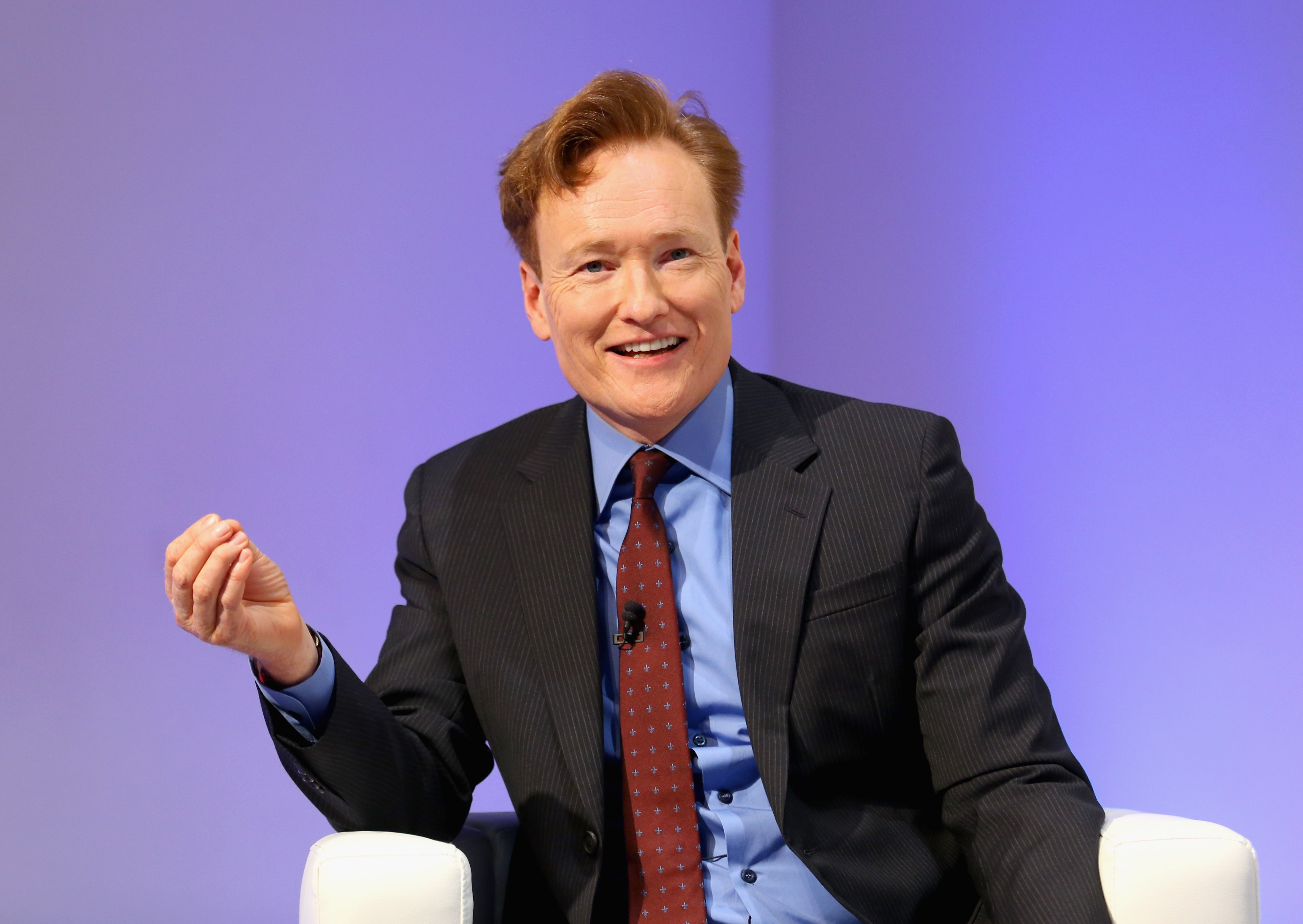Conan O'Brien talks and gestures with one hand. He is sitting in a chair and wearing a blue shirt, red tie, and black jacket. 
