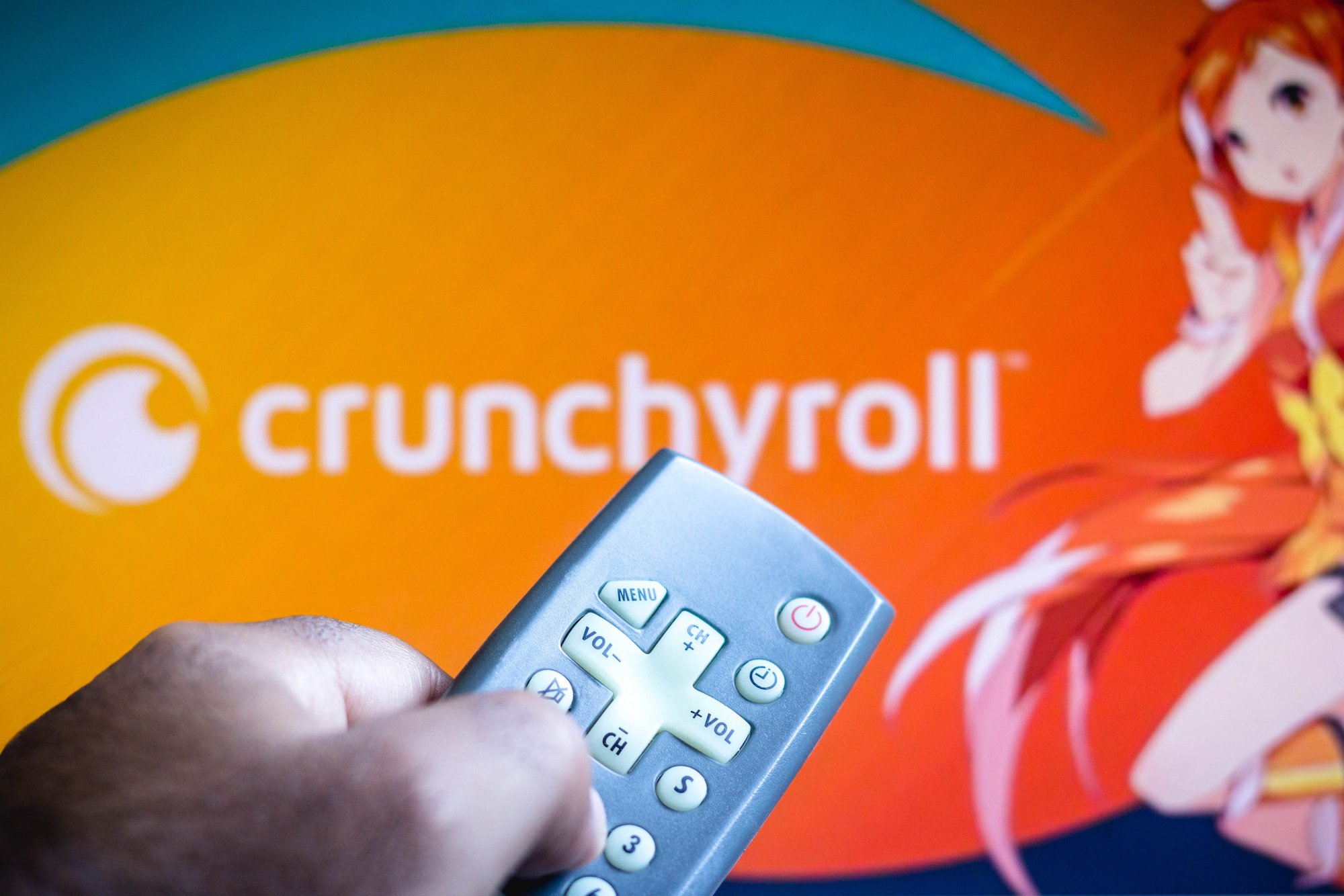 In this photo illustration a close-up of a hand holding a TV remote seen displayed in front of Crunchyroll logo