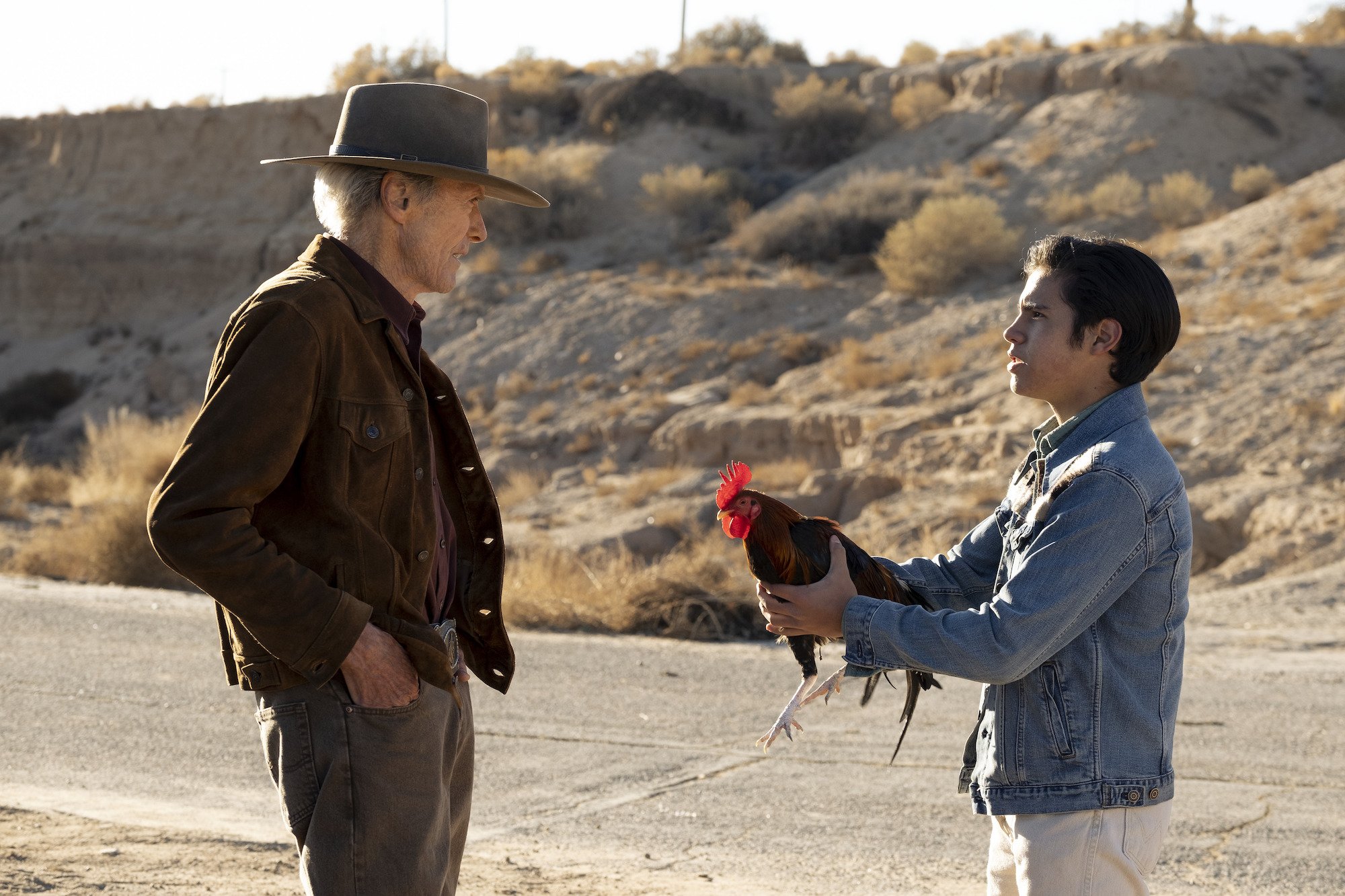 'Cry Macho': Eduardo Minett hands Clint Eastwood his rooster. Eastwood wears a brown cowboy hat and brown suede jacket with jeans. Minett wears a blue demin jacket and white jeans. They stand next to a barren road with dessert terrain surrounding them during the daytime.