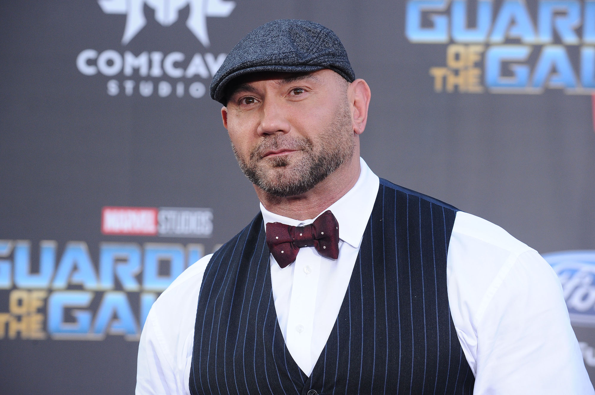 Marvel star Dave Bautista does not reprise his role in 'What If...?' In this photo, he's wearing an ivy cap, black striped vest, white shirt, and bowtie. He's looking past the camera and there's a 'Guardians of the Galaxy' wall behind him.