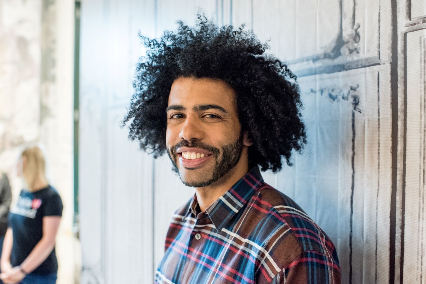 Daveed Diggs from 'Hamilton' wearing a blue, white, red, and brown shirt leaning against a light colored wall.