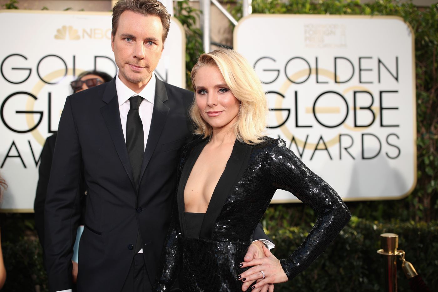 Dax Shepard dressed in a black suit with a white shirt and black tie with Kristen Bell dressed in a solid black dress in front of a Golden Globe Award sign.