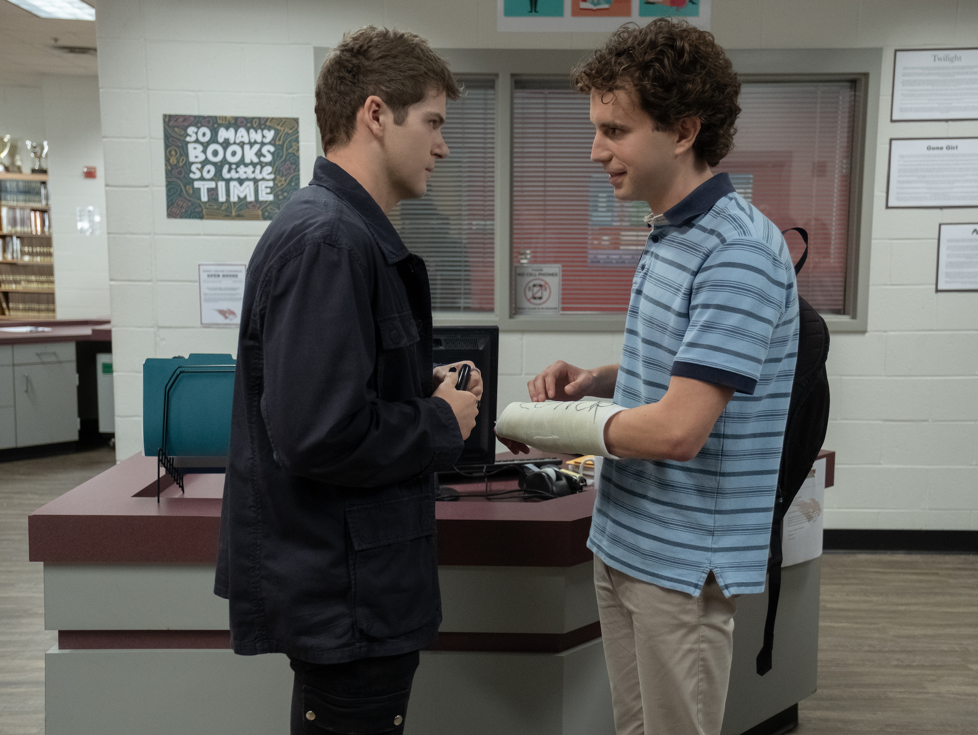 Colton Ryan and Ben Platt as Connor Murphy and Evan Hansen in the 'Dear Evan Hansen' movie. Ryan signs Platt's cast in a high school library. A desk with a computer and blue envelopes is behind them with a window looking out to a hallway. Platt's age in 'Dear Evan Hansen' has been a point of contention since the trailer came out, as people think he looks too old at 27 to be playing a high school student. Ryan is 26 and plays a character the same age as Platt's.